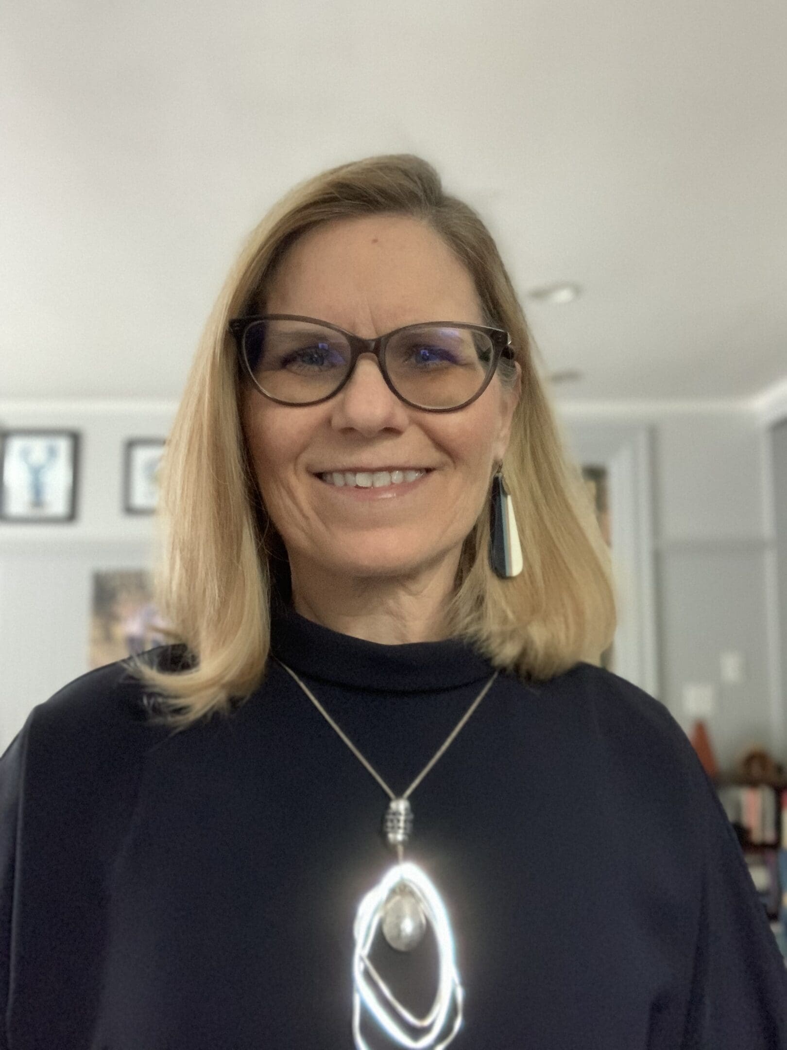 Smiling woman wearing glasses and a pendant necklace indoors.