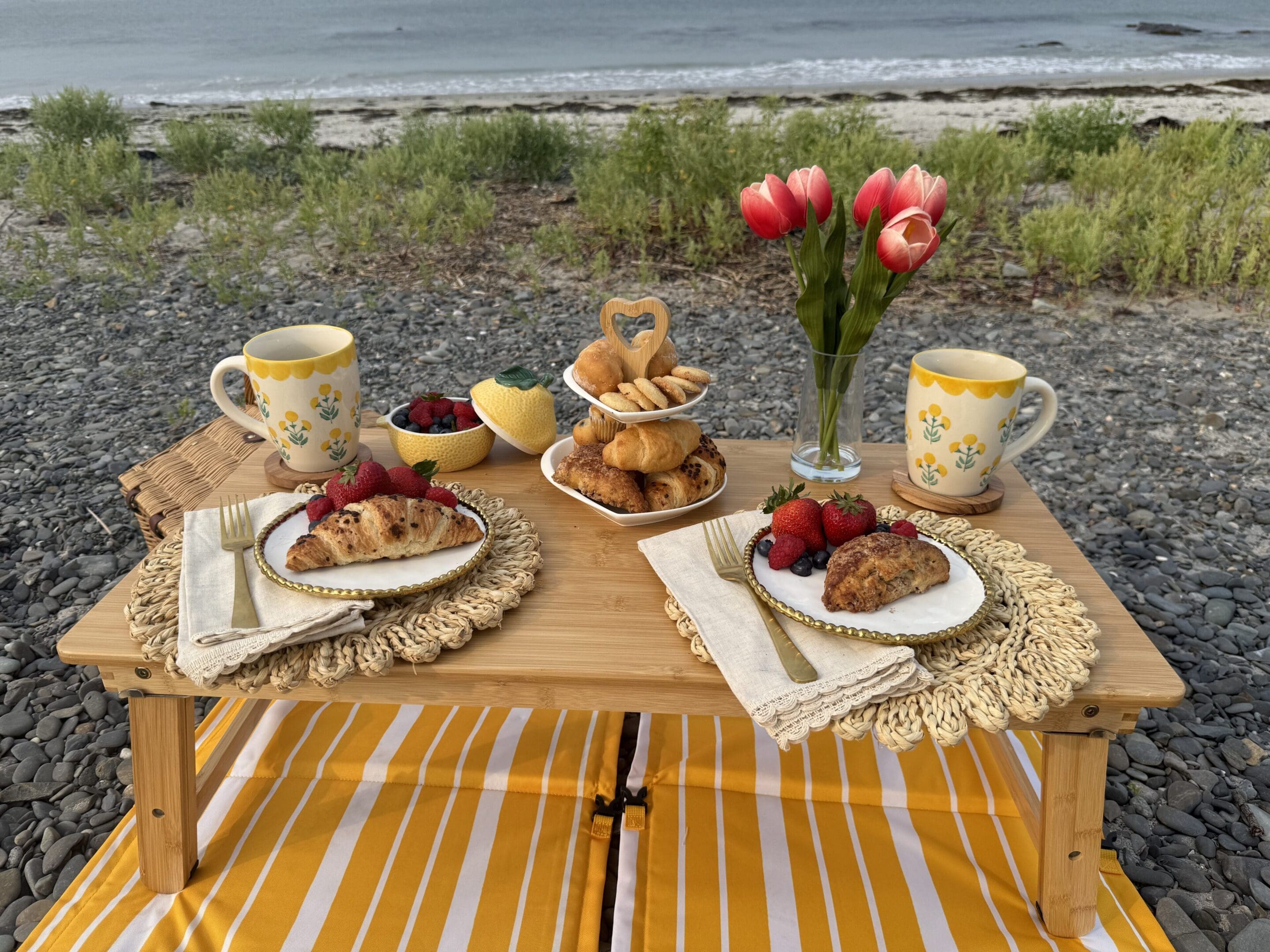 Cozy outdoor tea setup with pastries, tea cups, and flowers on a picnic blanket by the water.